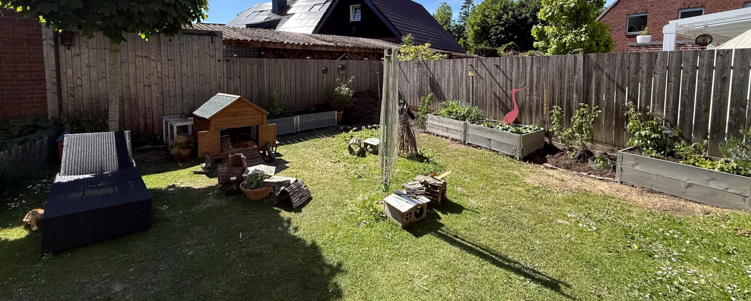 Panoramabild aus dem Garten mit Palettenbeeten, unterbrochen durch Sträucher. Im Vordergrund eine Liege und ein Meerschweinchenstall