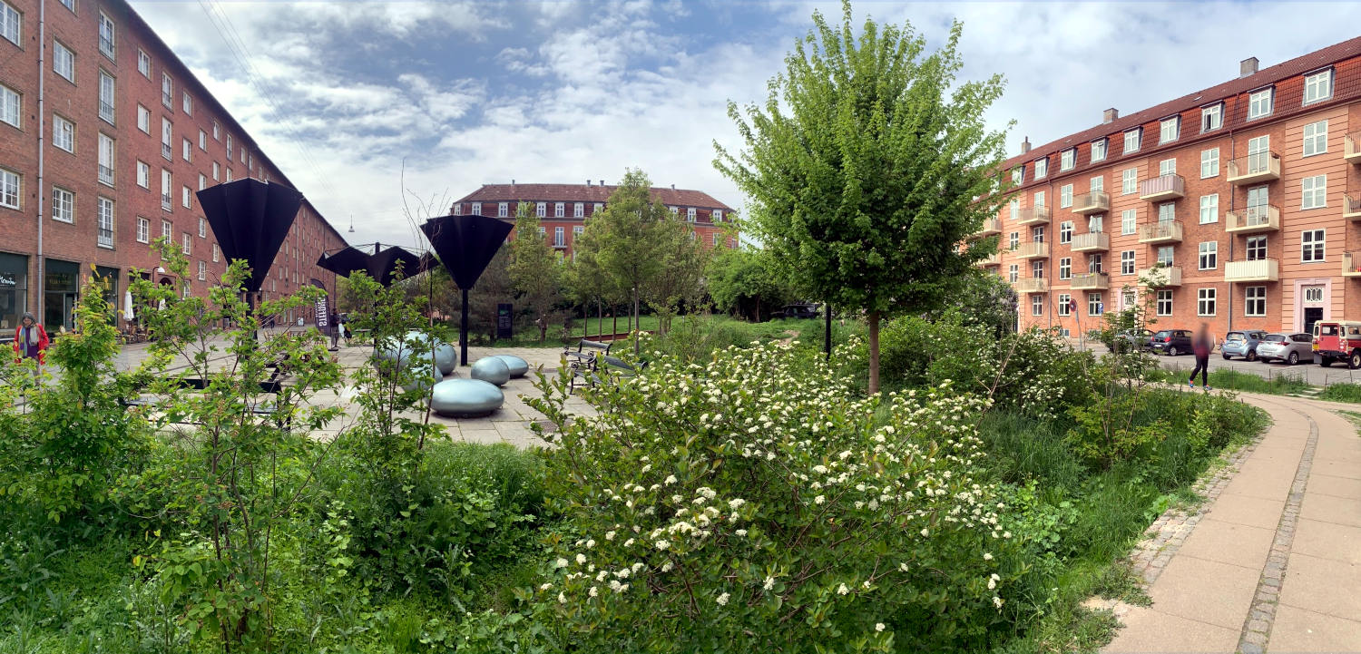 Ein Platz inmitten von Mehrfamilienhäusern. Es gibt viele Büsche, Wiese, Bäume und Metallkugeln zum Hinsetzen.