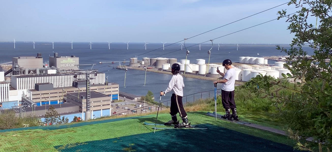 Blick vom Top der Müllhalde: Zwei Skifahrer stehen auf Skiern und Kunstrasen oben. Man blickt aufs Meer, auf den Hafen und auf Windkraftanlagen im Wasser.