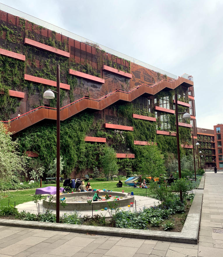 Parkhaus aus Metall, außen führt eine Metalltreppe aufs Dach hinauf. Aus der Fassade wächst Grünzeug. Davor ein Sandkasten, Eltern und Kindern