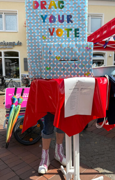 Gebastelte Wahlkabine auf einem Stehtisch, darauf die Buchstaben "Drag your vote". Es sind Füße zu sehen. Jemand "wählt" gerade.