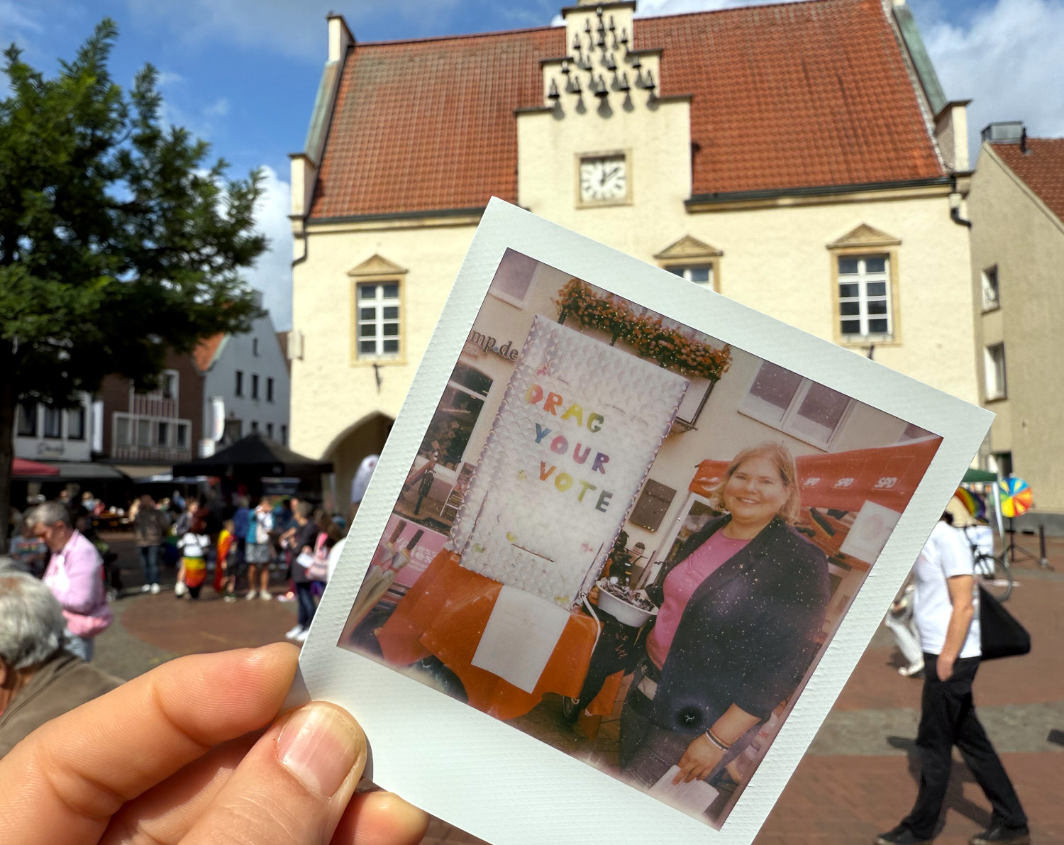 Ein Polaroidbild von Vanessa mit ihrer Wahlkabine. Eine Hand hält es in die Luft. Dahinter ist ein historisches Rathaus mit Glockenspiel zu sehen.