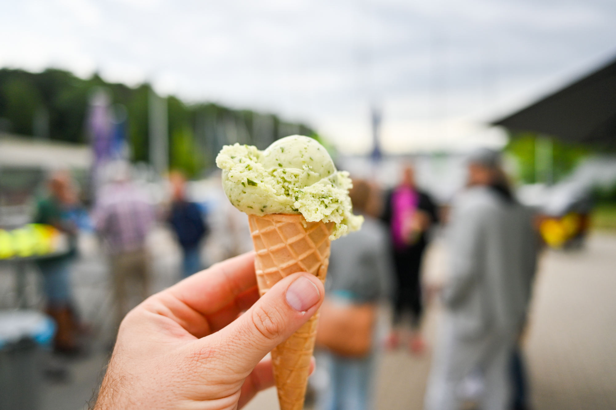 Jemand hält eine grünliche Kugel Eis hoch, dahinter Menschen, Wasser und Bootsmasten