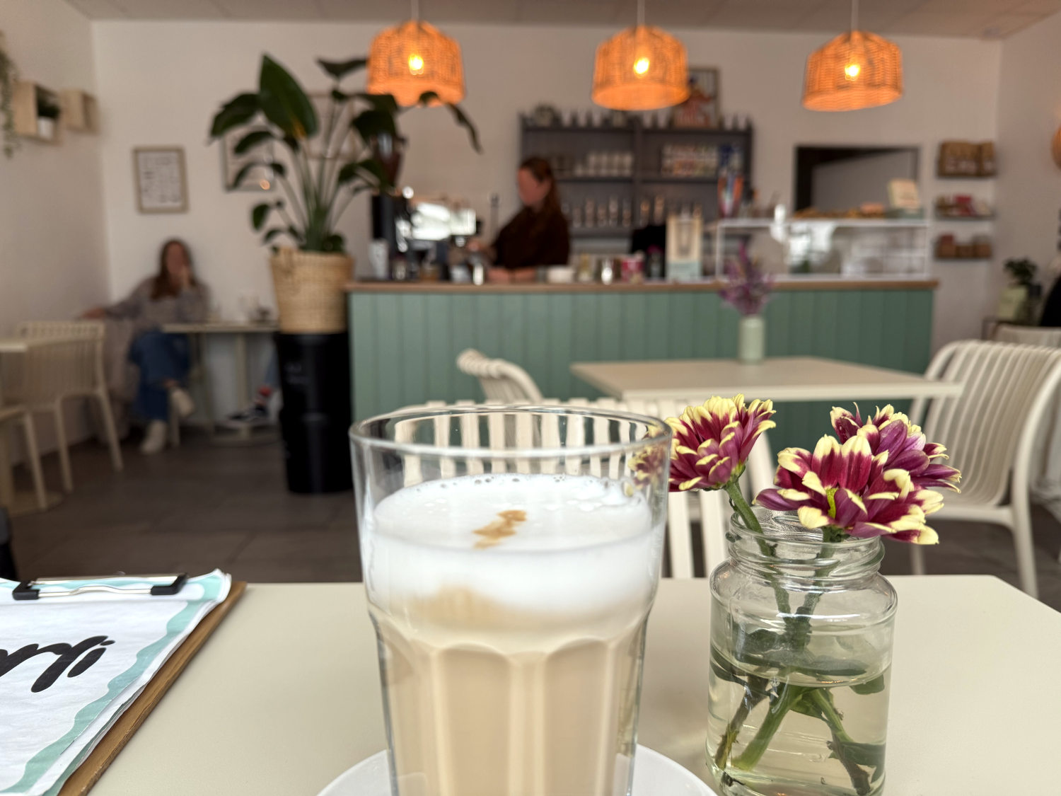 Im Vordergrund ein Tisch mit einer Latte Macchiato und Blumen in einem Glas, im Hintergrund eine Theke mit Lampen darüber und zwei Frauen. Die Atmosphäre ist anheimelnd.