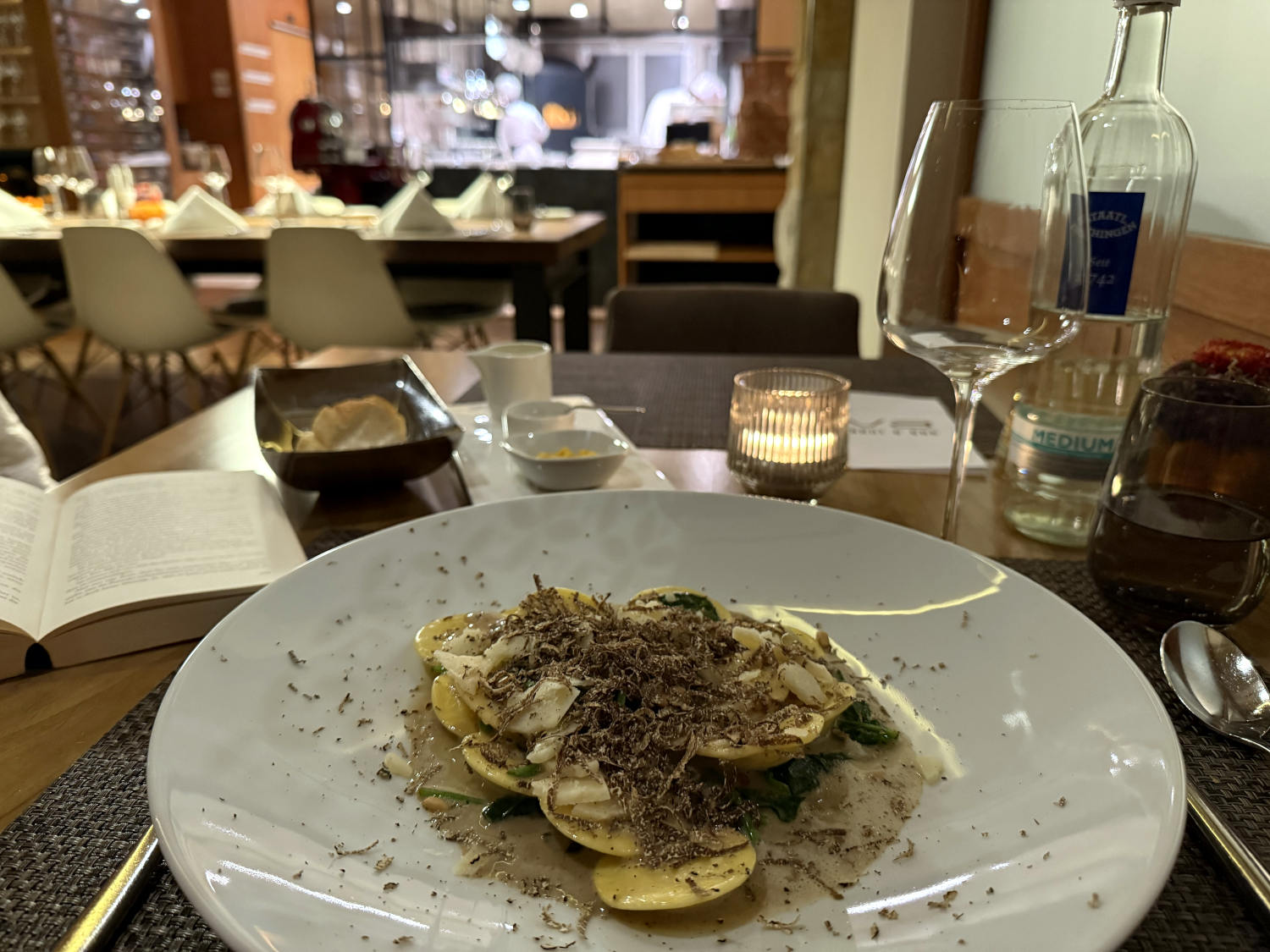 Ein Teller mit Ravioli, reicht bedeckt mit Trüffel. Gedämpftes Licht, ein Weinglas, eine Wasserflasche, ein Buch, im Hintergrund Restaurant.
