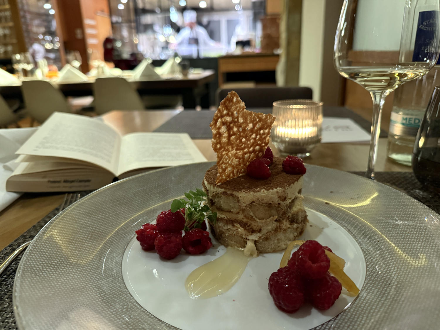 Ein rund ausgestochenes Stück Tiramisu, darin steht etwas Karamallisiertes, daneben Himberren. Daneben ein Weinglas und ein Buch. Im Hintergrund Köche hinter einer Theke.