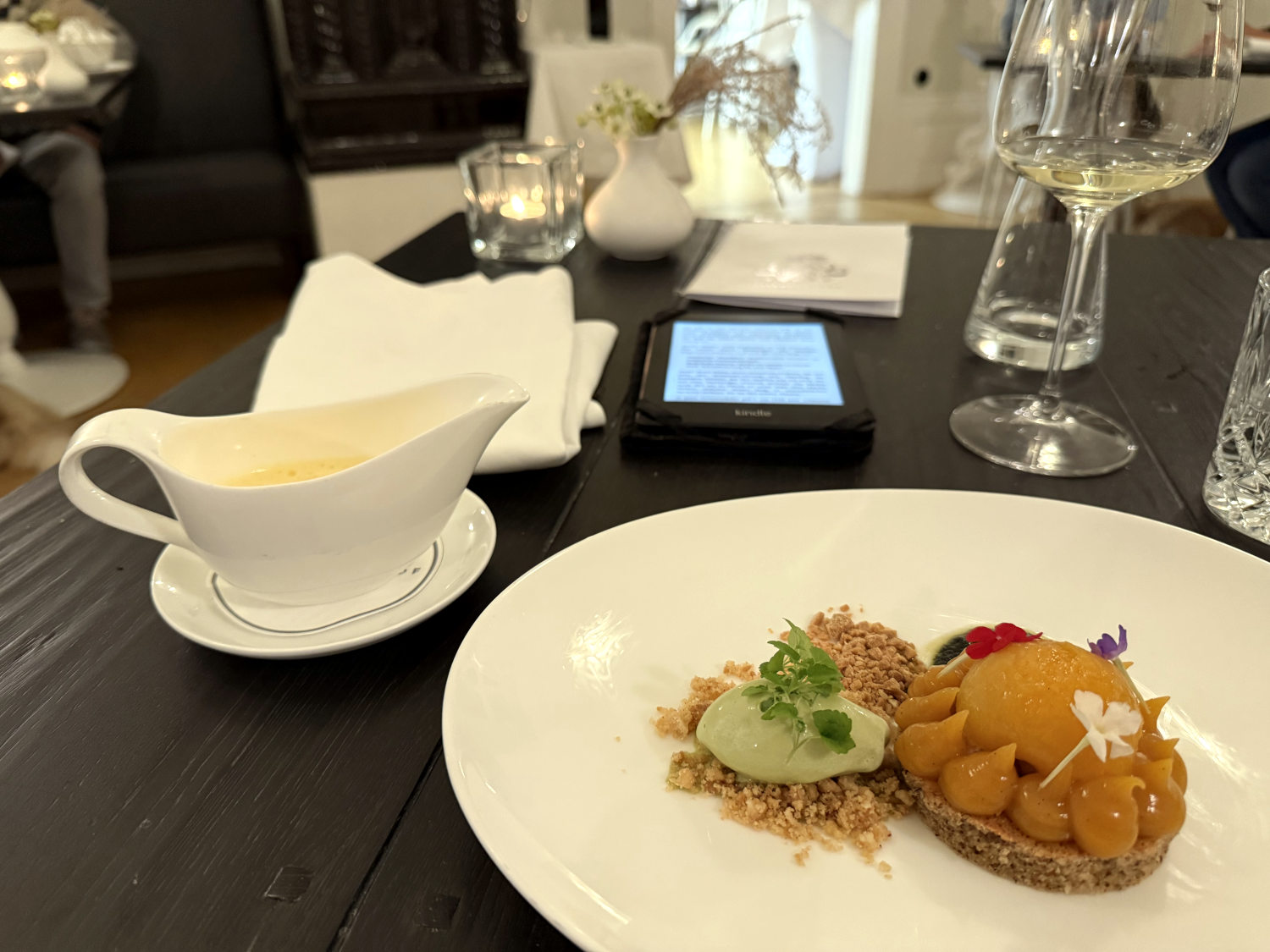 Ein Aprikosentörtchen mit Pistazieneis, daneben eine Sauciere mit Champagnerschaum, auf dem Tisch ein Glas Wein, eine Kerze und ein Kindle.