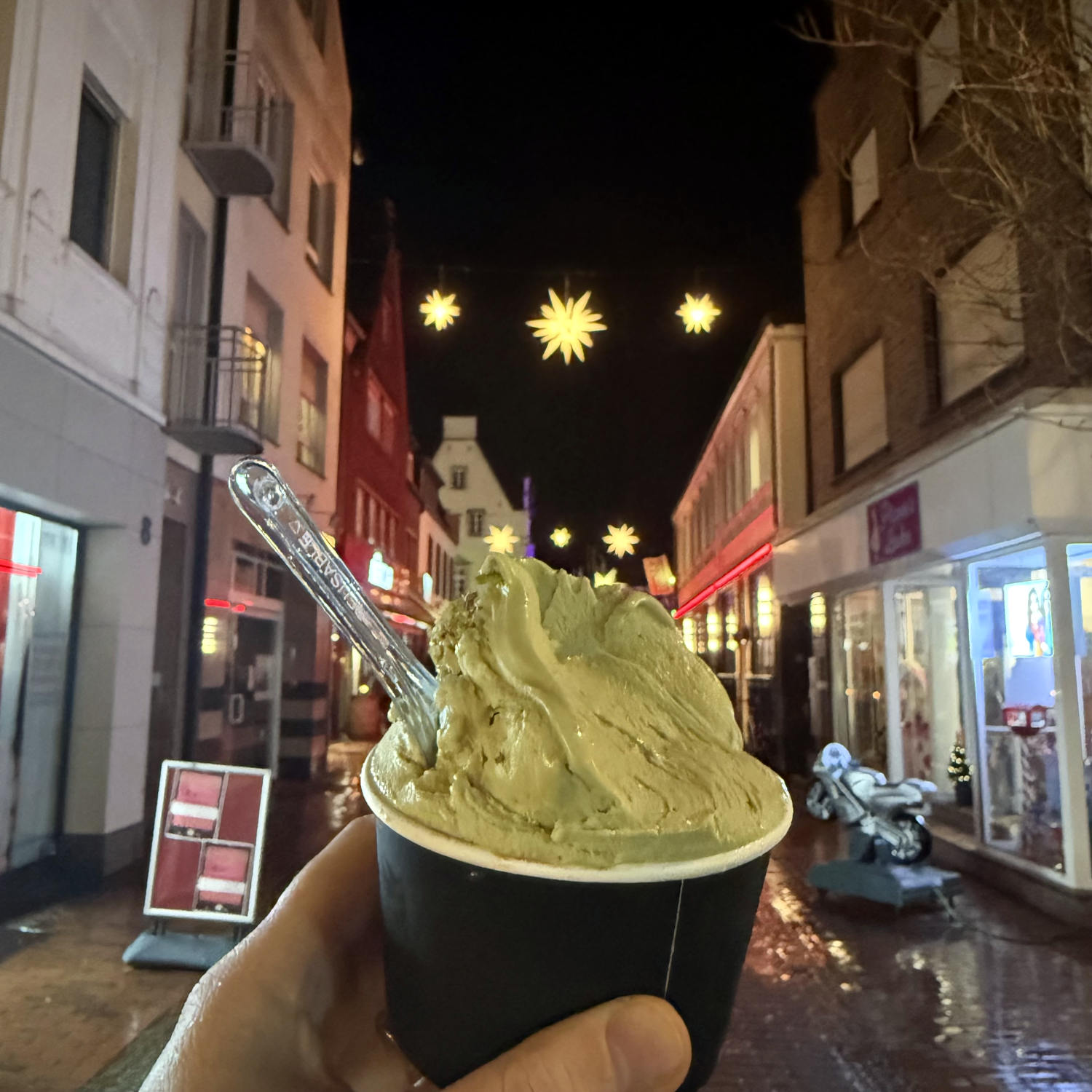 Eine Halt hälte einen Eisbecker mit Pistazienreis. Im Hintergrund eine verregnete Straße und Sterne zwischen den Häusern.