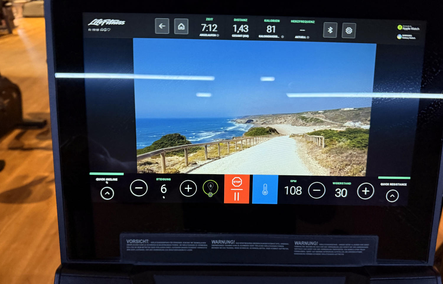 Bildschirm eines Fitnessgeräts. Darauf das Bild eines sonnigen Küstenwegs, in der Ferne das Meer.