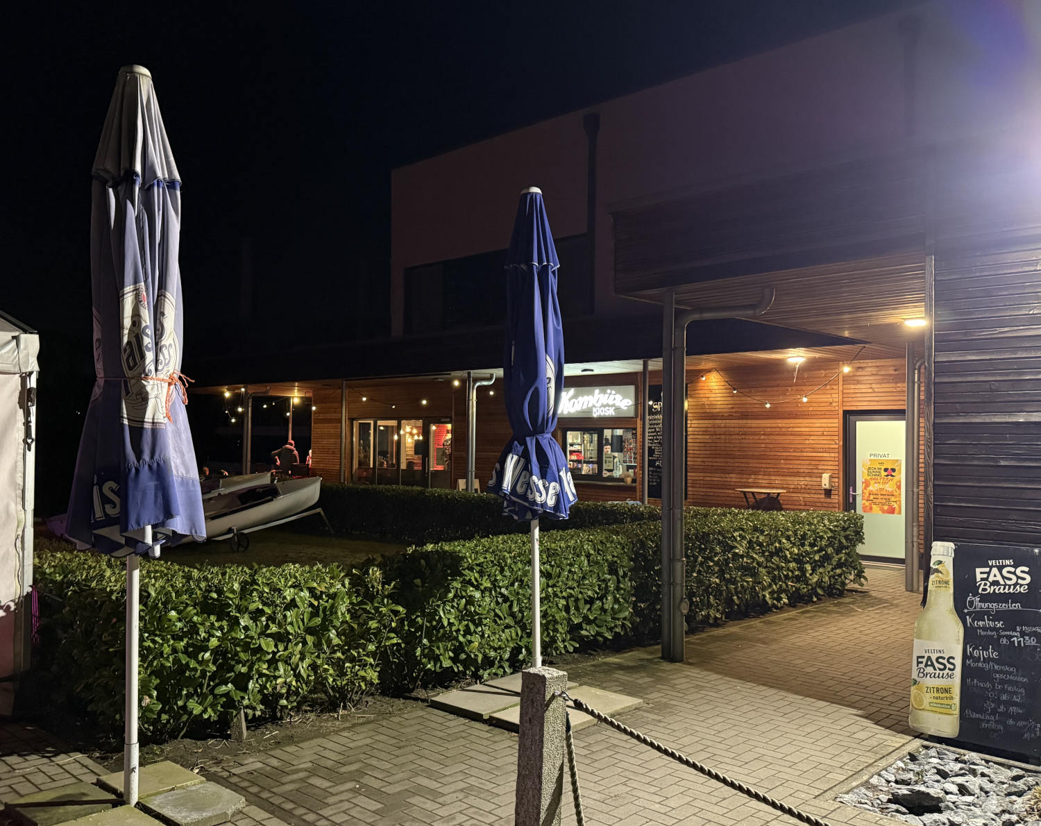 EIngang zum Biergarten, beleuchtet bei abendlicher Dunkelheit, ein Holzbäude, davor Schirme, eine Tafel mit Angeboten, es deutet sich in der Dunkelheit ein See mit Booten an