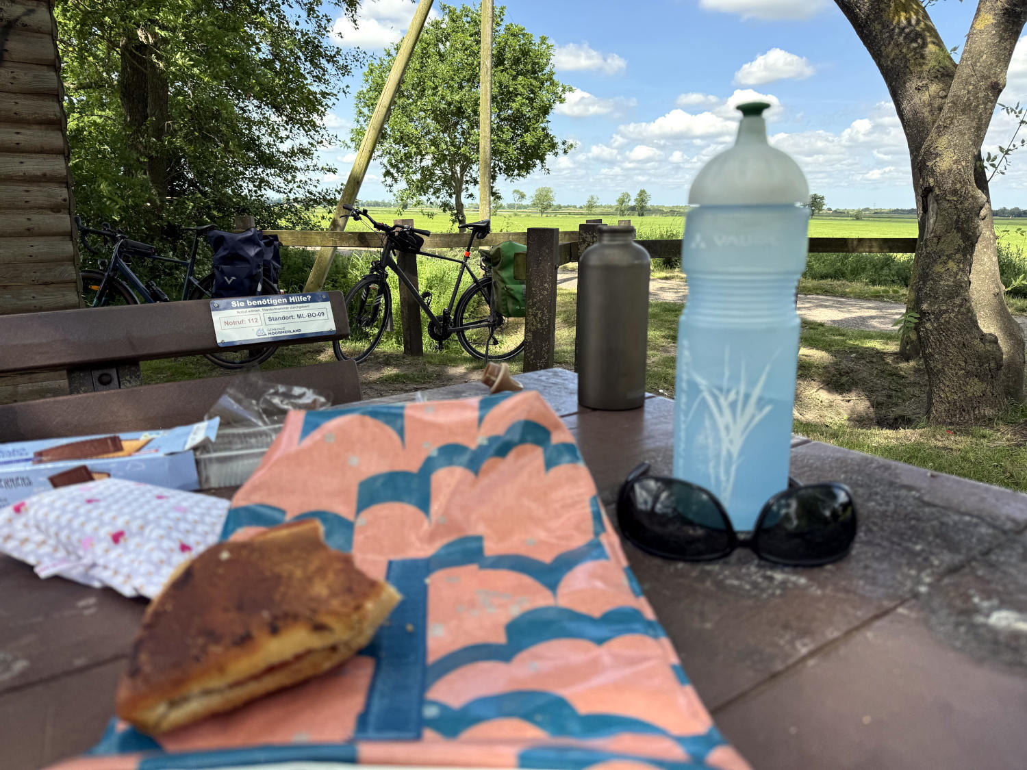 Im Vordergrund, unscharf, ein Beutel, darauf ein angebissenes Brötchen, eine Trinkflasche, Kekse und eine Sonnenbrille. Im Hintergrund Fahrräder, Wiese, Sonnenschein.
