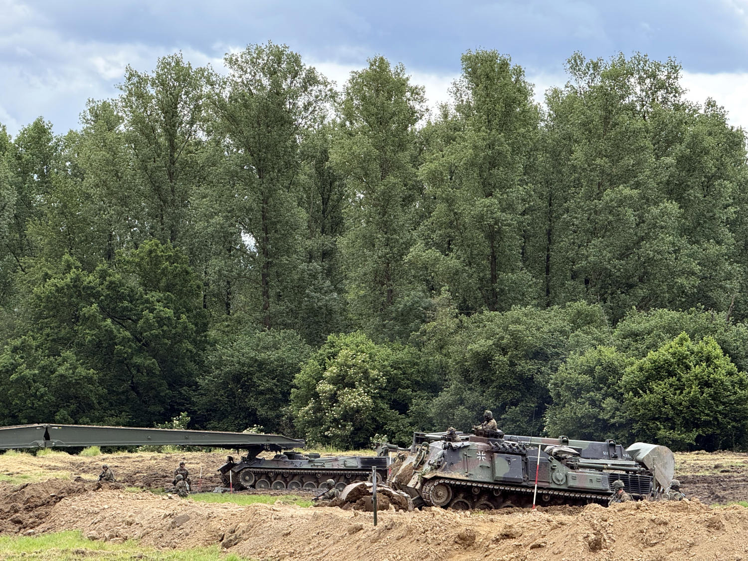 Ein Panzer, der baggern kann ("Dachs"), und ein Brückenpanzer in einem Feld, das schon ziemlich durchwühlt ist. Im Hintergrund Bäume. 