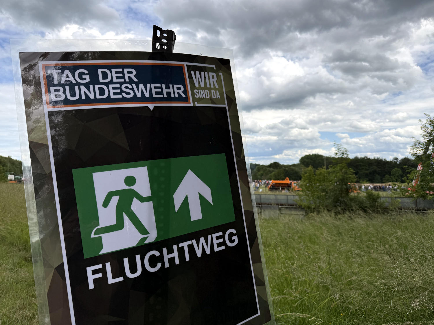 Schild vor einer Wiese, im Hintergrund sieht man eine Menschenmenge: "Tag der Bundeswehr" - "Fluchtweg". Oben rechts der Spruch: "Wir sind da."
