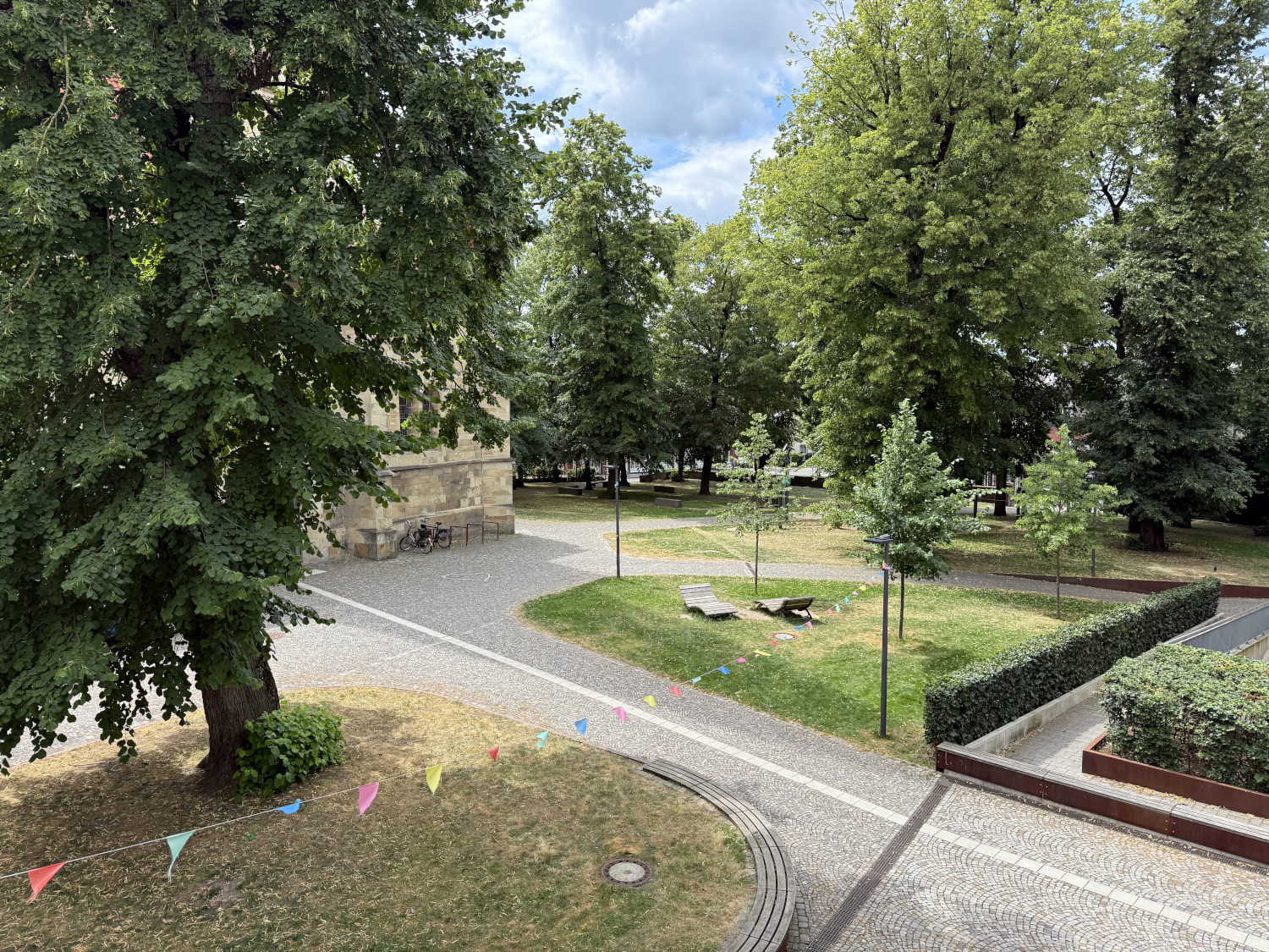Blick von oben in einen Kirchhof mit Bäumen, Bänken und Rasen. Bunte Fähnchen sind zwischen zwei Laternen gespannt.