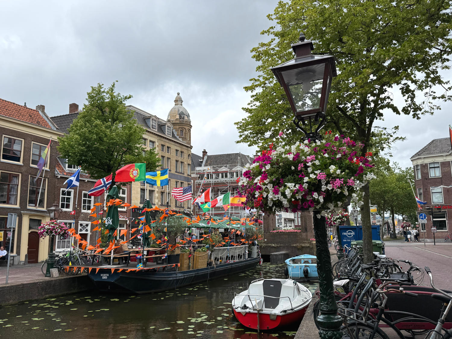 Gracht in Leiden. Ein Boot mit Gastronomie und wehenden Fahnen ist am anderen ufer vertäut. Im Vordergrund ene Laterne mit Geranien.