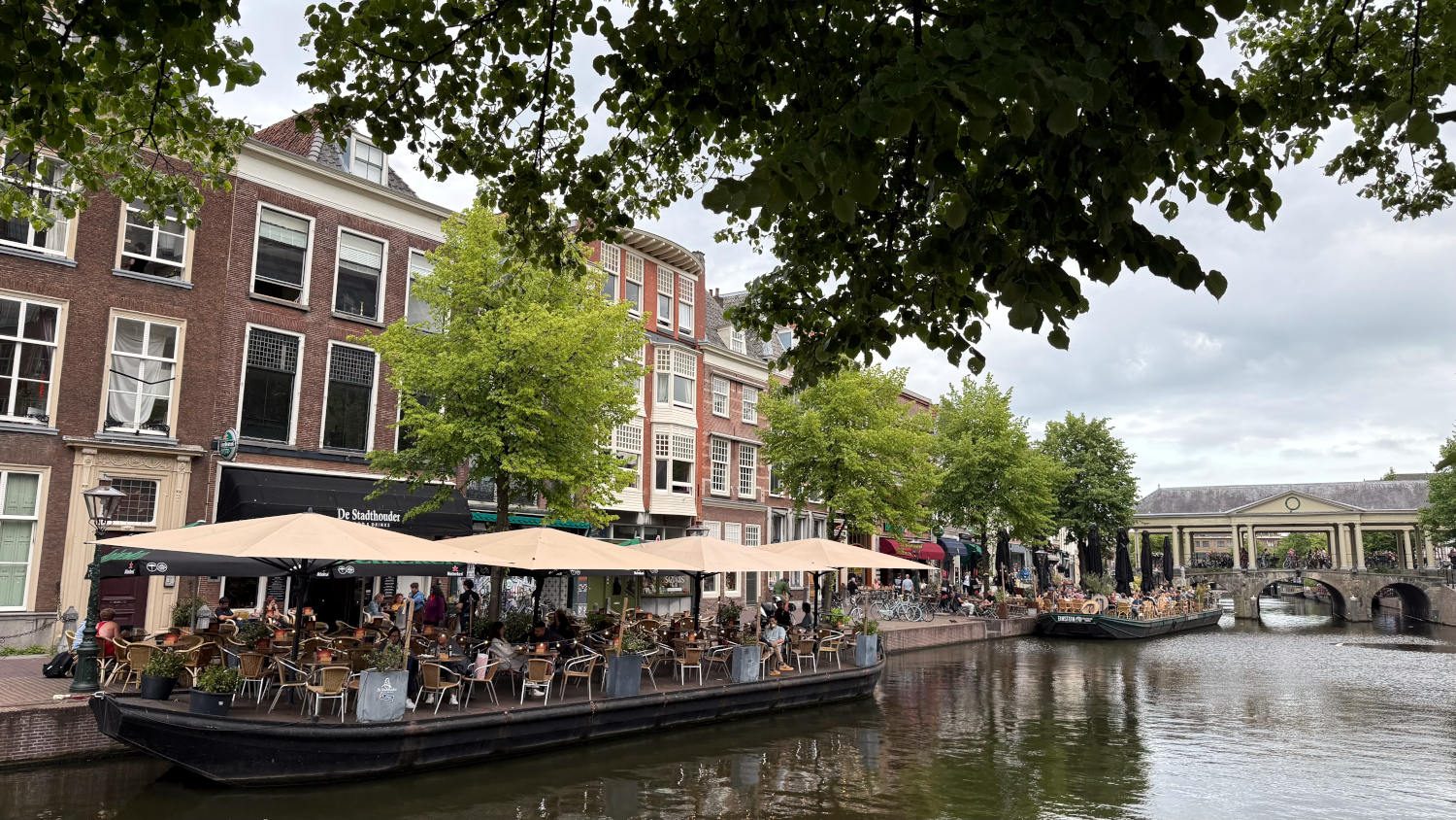Gracht in Leiden, im Hintergrund eine Brücke. Im Vordergrund ein Schiff mit Gastronomie. Die Sonnenschirme sind geöffnet, Menschen sitzen auf Stühlen.