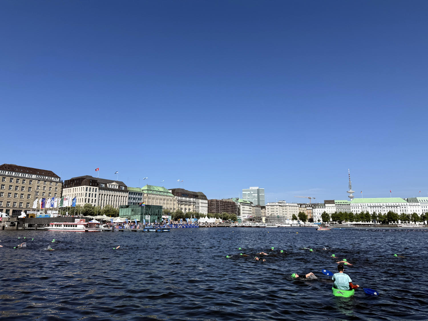 Binnenalster. Es schwimmen Leute mit farbigen Badekappen darin. Ein Mensch in einem Kajak sorgt für Sicherheit.