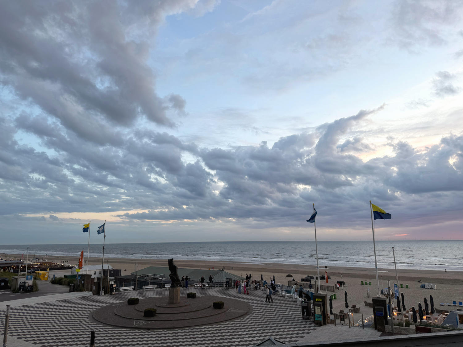 Blick auf Strand und Meer mit wolkigem Abendhimmel. Im Vordergrund ein Platz mit einer Figur. Es ist windig. Auf dem Strand stehen Büdchen und Strandkörbe.