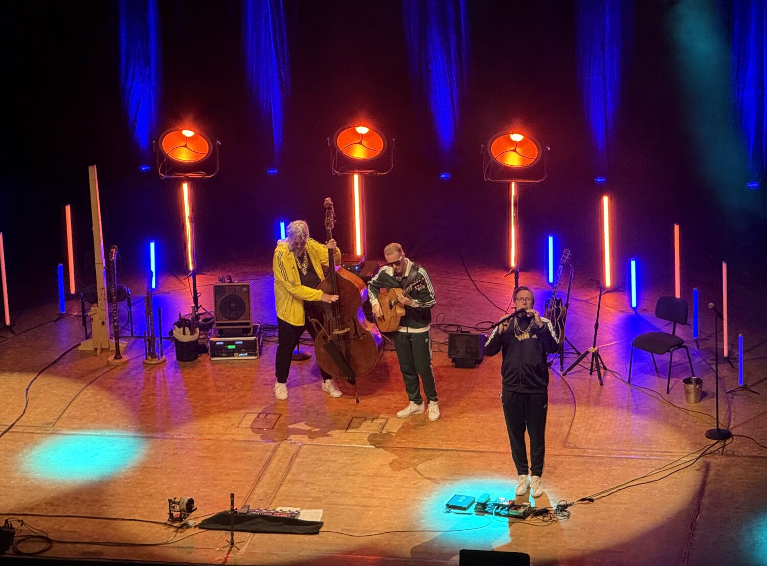 Drei Männer auf der Bühne mit Kontrabass, Gitarre und zwei (!) Blockflöten im Mund. Alle drei tragen Trainingsanzüge und Goldkettchen.
