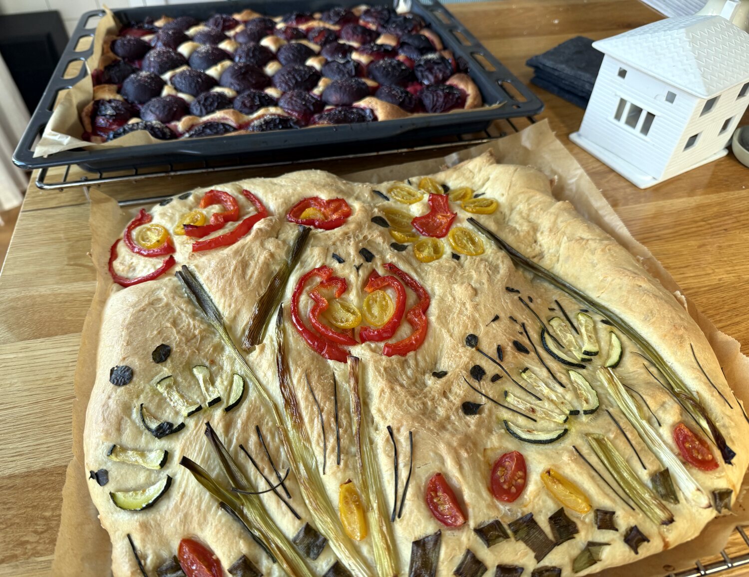 Foccaccia auf dem Blech, Gemüse bildet eine Blumenwiese. Im Hintergrund ein Blech Pflaumenkuchen.