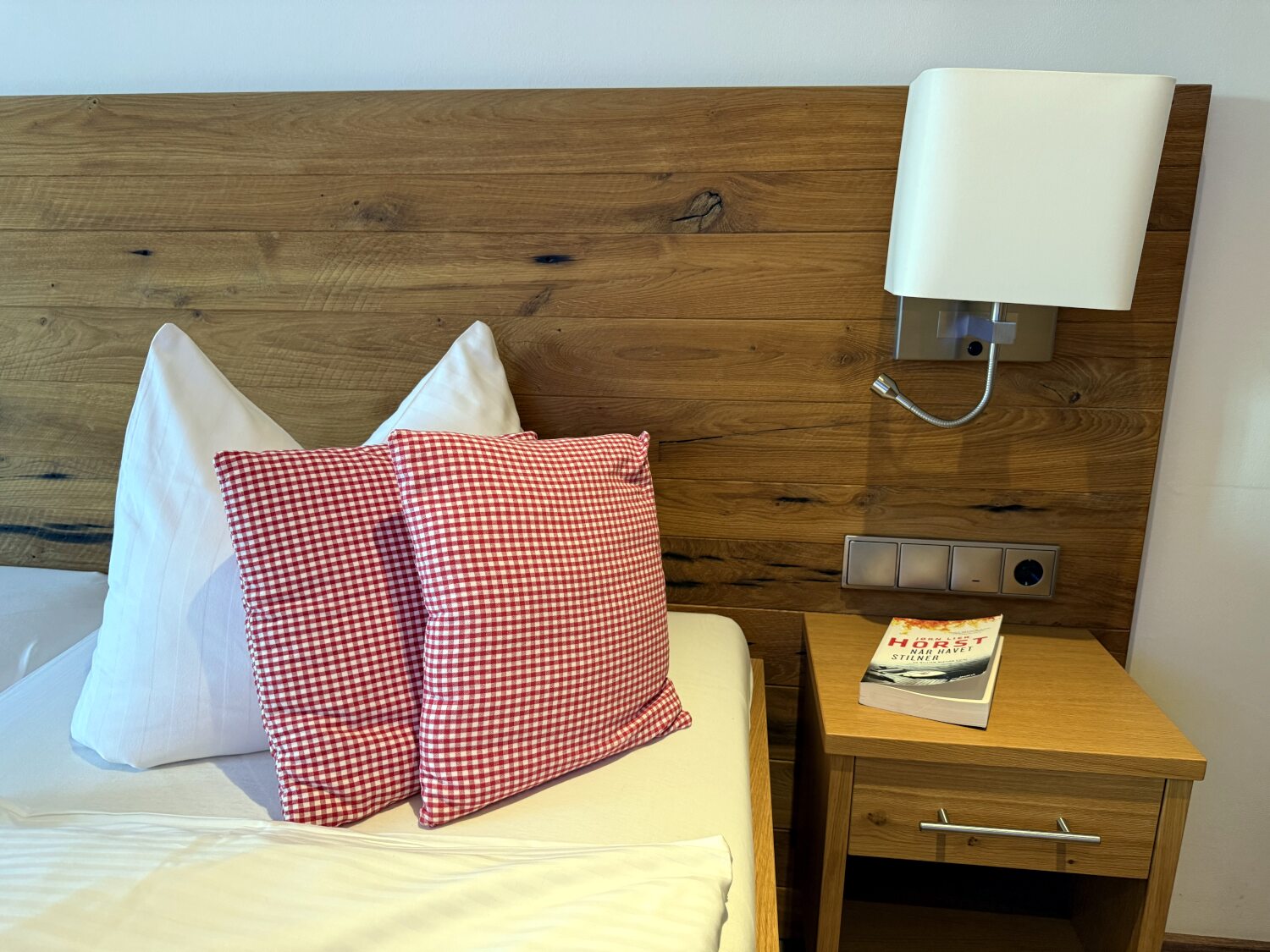 Hotelbett aus Holz mit karierten Kissen. Auf dem Nachttisch ein Buch.